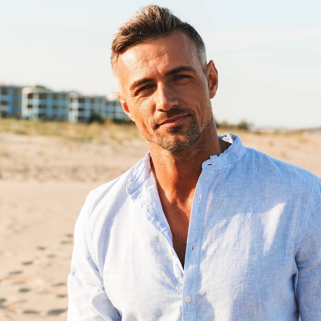 Fit, athletic man in a light blue linen shirt with a sharp, youthful jawline standing outdoors on a sunny beach.